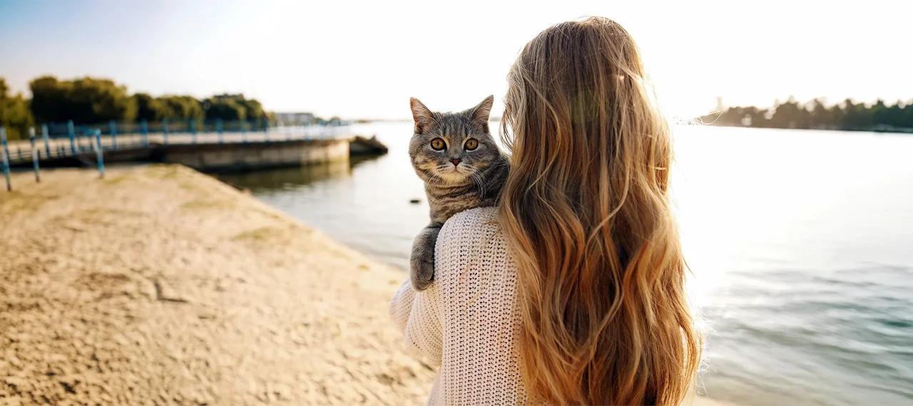 A woman holds her gray cat while walking by a lake